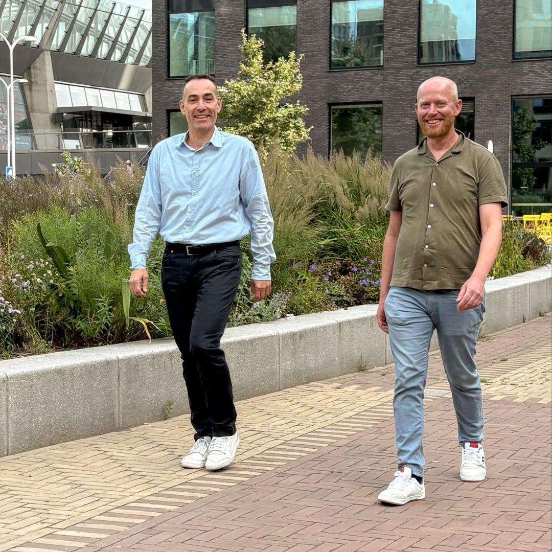 Twee mannen lopen buiten langs een modern kantoorgebouw met grote ramen en balkonnetjes. Ze kijken ontspannen en lachend in de richting van de camera.