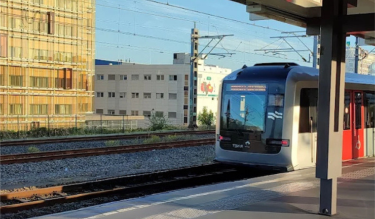 Een metrotrein met rode en grijze deuren staat op een modern perron, met gebouwen en spoorlijnen zichtbaar in de achtergrond.