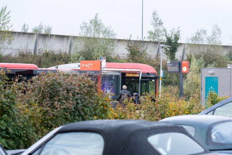 Een reiziger stapt in een rode R-net bus bij een bushalte op een P+R-terrein, omringd door struiken en geparkeerde auto’s.